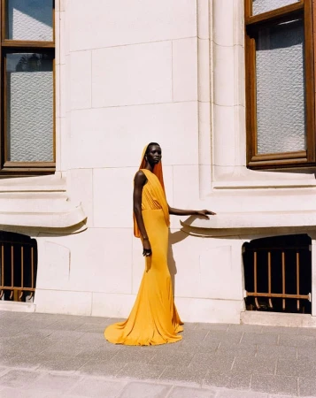 Elegant black female fashion model in a long, draped, mustard-yellow gown posing against an architectural building with windows.
