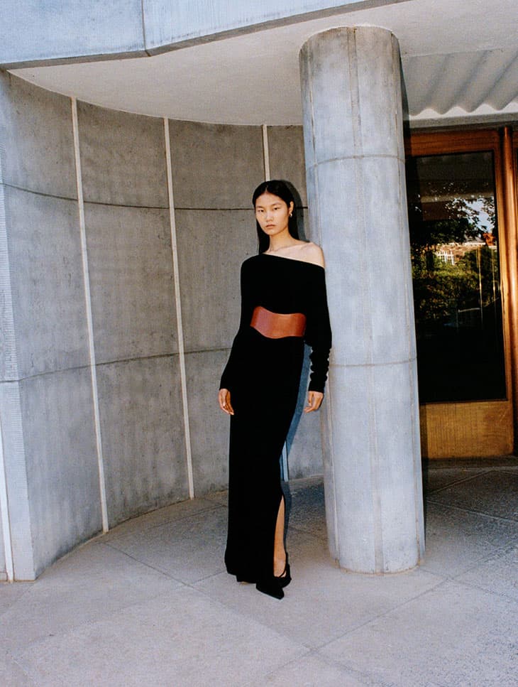 Elegant Asian woman poses in a black dress with a brown belt against a concrete wall in a fashion editorial shot.