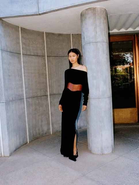 Elegant Asian woman poses in a black dress with a brown belt against a concrete wall in a fashion editorial shot.