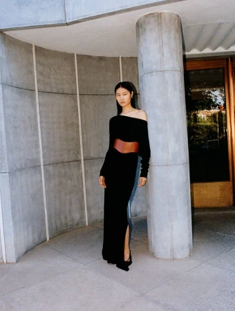 Elegant Asian woman poses in a black dress with a brown belt against a concrete wall in a fashion editorial shot.