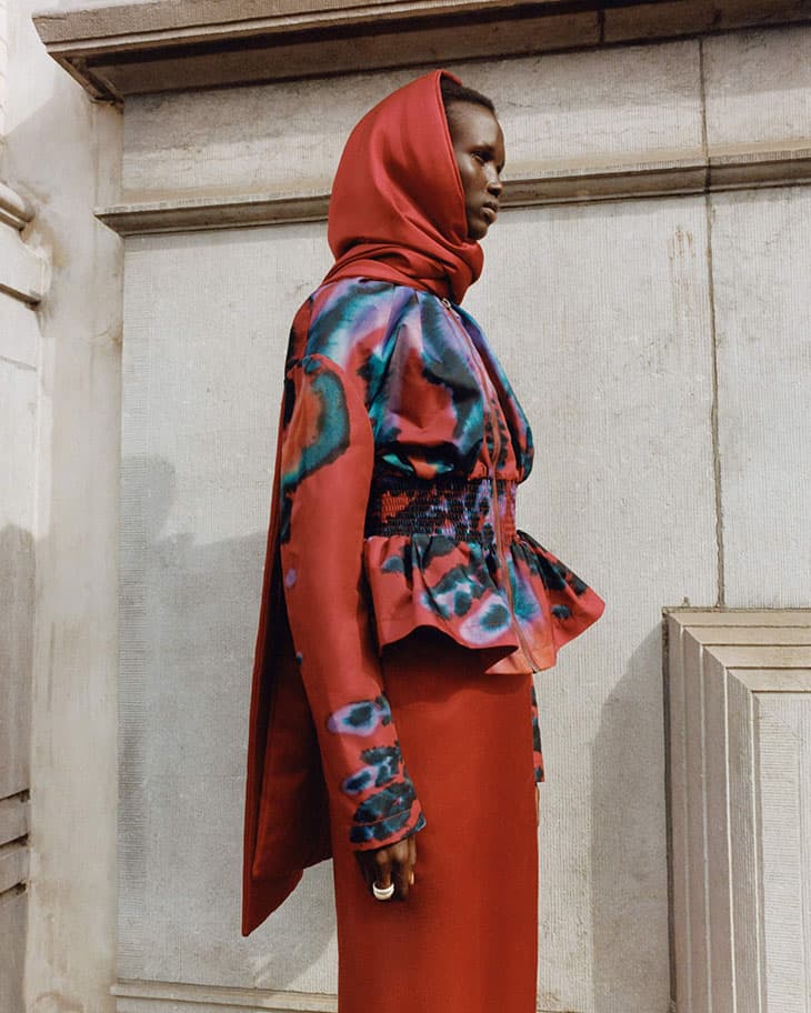 Woman wearing an orange headscarf and colorfully patterned dress stands in front of a concrete wall with architectural trim details.