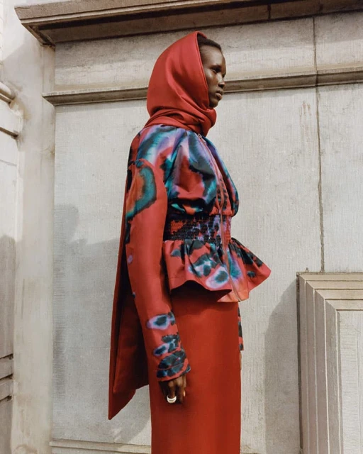 Woman wearing an orange headscarf and colorfully patterned dress stands in front of a concrete wall with architectural trim details.
