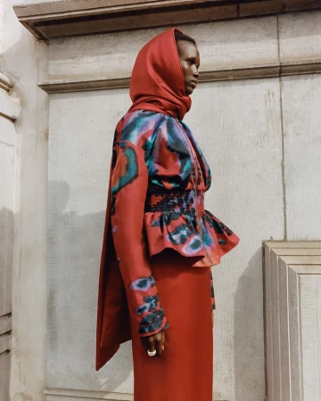 Woman wearing an orange headscarf and colorfully patterned dress stands in front of a concrete wall with architectural trim details.