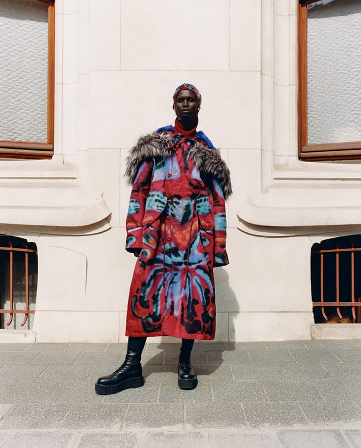 A person standing outdoors on a sidewalk, wearing a colorful coat with a fur collar and black boots, posing for a fashion editorial.