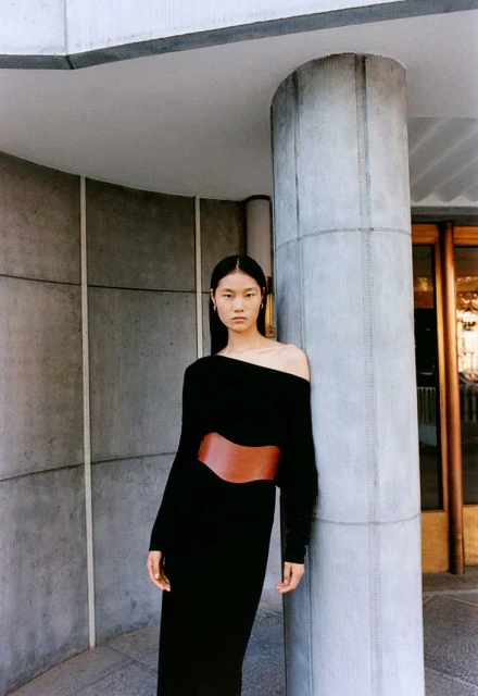 Asian woman wearing black off-the-shoulder dress with wide brown belt stands near a gray concrete pillar and arched wall indoors.