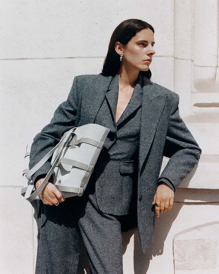 A woman in a grey suit holds a light grey leather handbag, posing against a textured, light-colored wall in an outdoor setting with natural light.