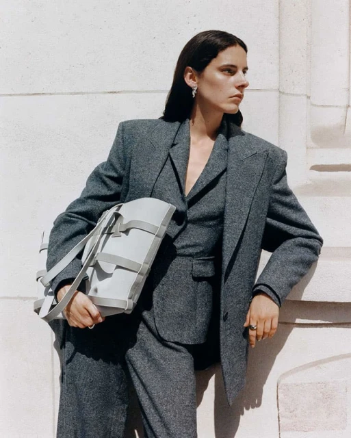 A woman in a grey suit holds a light grey leather handbag, posing against a textured, light-colored wall in an outdoor setting with natural light.