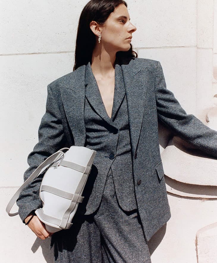 Adult woman in a grey suit with shoulder bag standing in an indoor environment with natural lighting and a neutral-colored wall.
