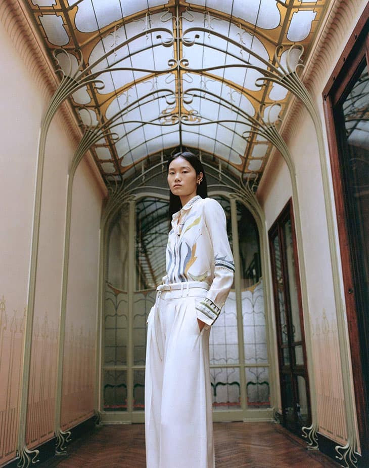 An Asian woman in a white patterned shirt and wide-leg pants stands in a hallway with ornate architectural details and natural light.