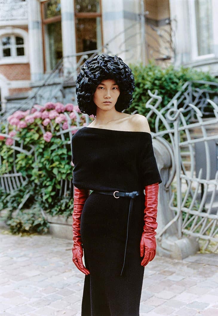 Asian woman in off-shoulder black dress with long red gloves and sculptural black headpiece poses outdoors near greenery and building.