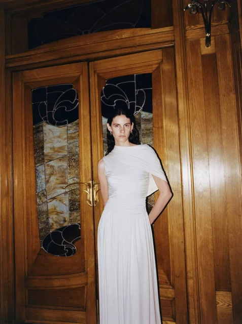 A woman stands in a white gown in front of wooden doors with stained glass, in an indoor environment with warm lighting.