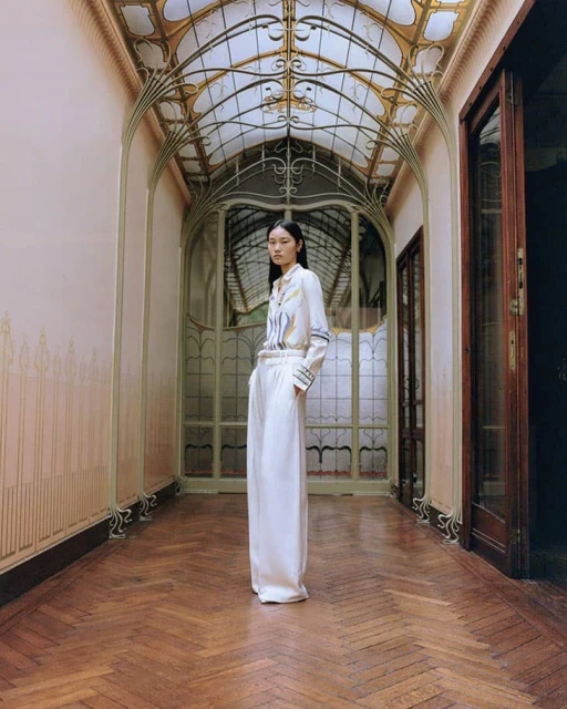 A stylish Asian woman stands in an ornate corridor wearing a white blouse and white palazzo pants in a fashion-forward pose, indoors.