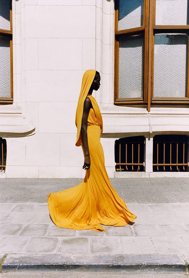 Profile shot of an adult female wearing a yellow hooded gown, standing on a stone walkway in front of a cream-colored building.