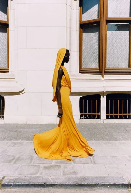 Profile shot of an adult female wearing a yellow hooded gown, standing on a stone walkway in front of a cream-colored building.