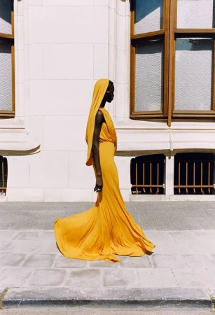Profile shot of an adult female wearing a yellow hooded gown, standing on a stone walkway in front of a cream-colored building.