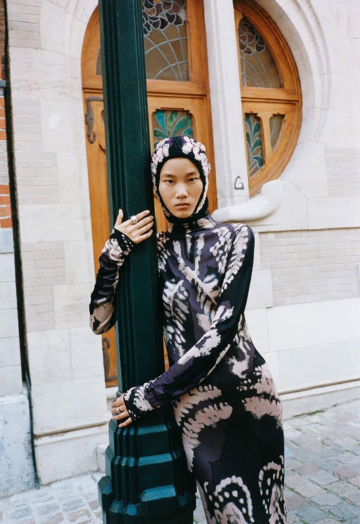 Asian woman in patterned dress and floral head covering, leans against a green pole with an urban background, fashion, and lifestyle.