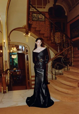 A fashionable adult woman stands in a hallway wearing a long black off-shoulder dress beside a wooden staircase and ornate design details.