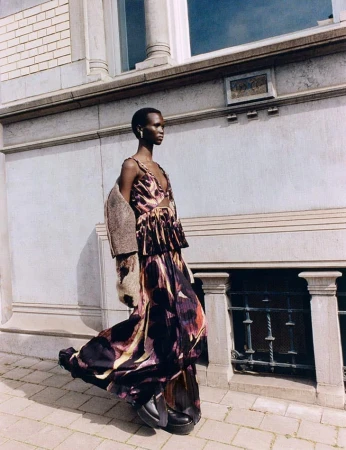 A solo adult female model poses in a colorful, flowing dress and black platform shoes on an outdoor city sidewalk.