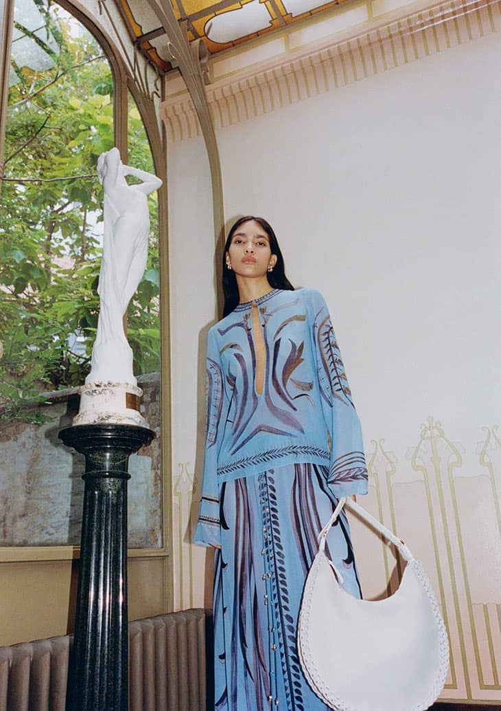 Indoor fashion shot of a woman in a blue patterned dress, holding a white bag, posing near a sculpture and arched window.