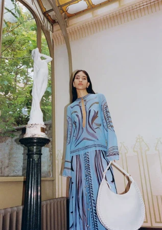 Indoor fashion shot of a woman in a blue patterned dress, holding a white bag, posing near a sculpture and arched window.