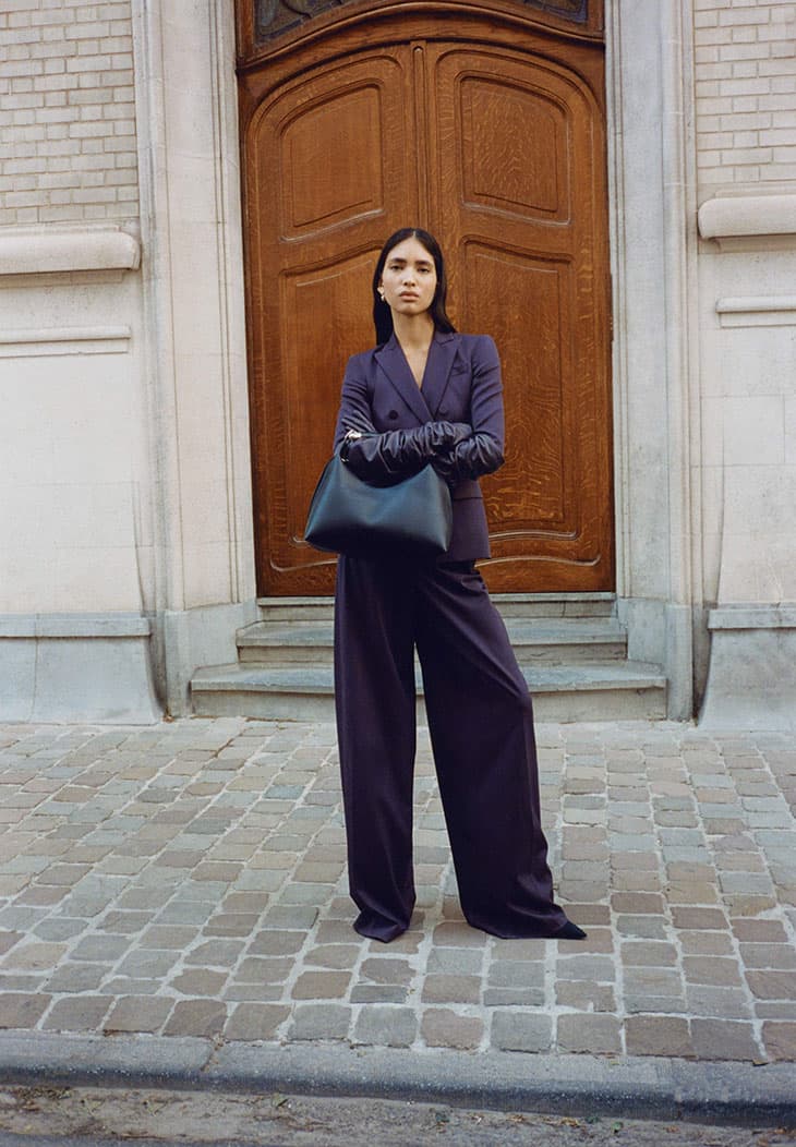Elegant brunette woman in a purple suit and long gloves stands outdoors holding a black tote bag with a wooden door behind her.