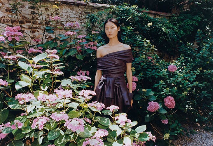 Model Canlan Wang stands among pink hydrangeas wearing an off-the-shoulder draped plum leather dress in a garden setting.