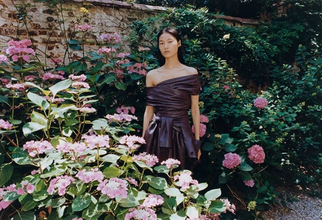 Model Canlan Wang stands among pink hydrangeas wearing an off-the-shoulder draped plum leather dress in a garden setting.