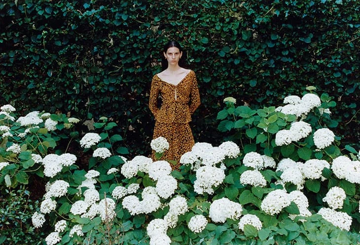 Adult woman stands in an outdoor garden with white flowers and a leafy green background, wearing a brown animal-print dress in daytime.