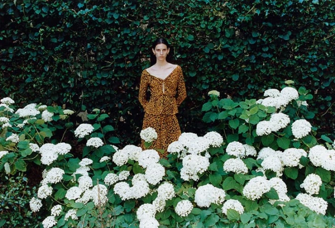 Adult woman stands in an outdoor garden with white flowers and a leafy green background, wearing a brown animal-print dress in daytime.