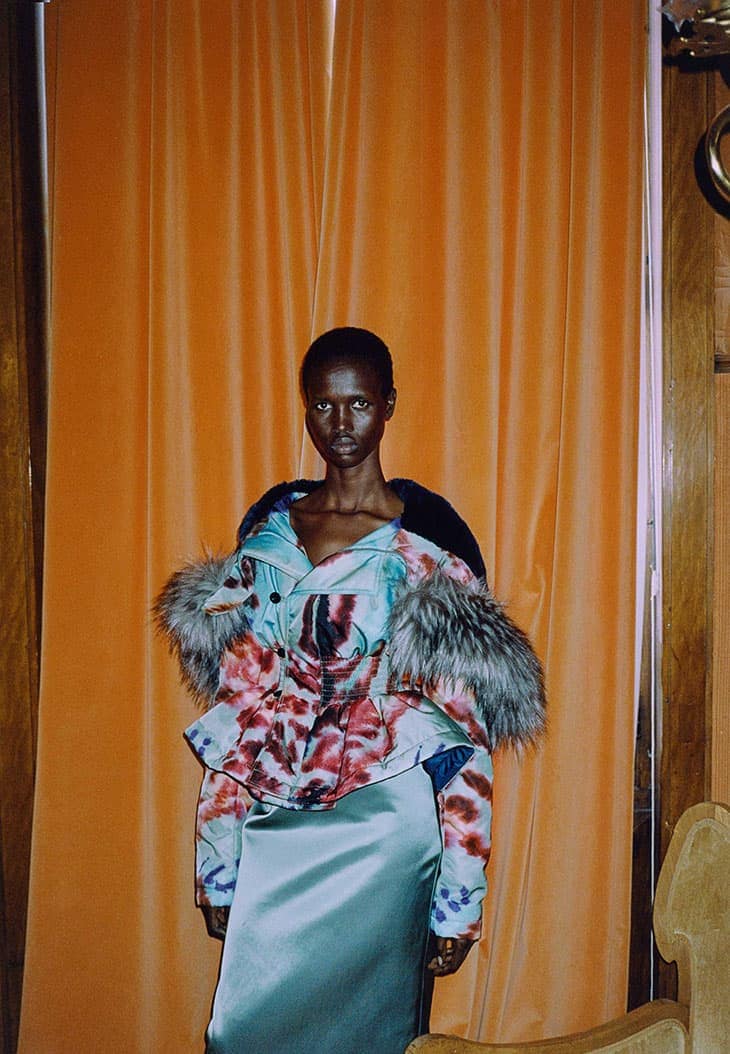 A statuesque Black woman poses indoors, wearing a floral jacket with fur trim and a teal skirt, standing against an orange curtain backdrop.