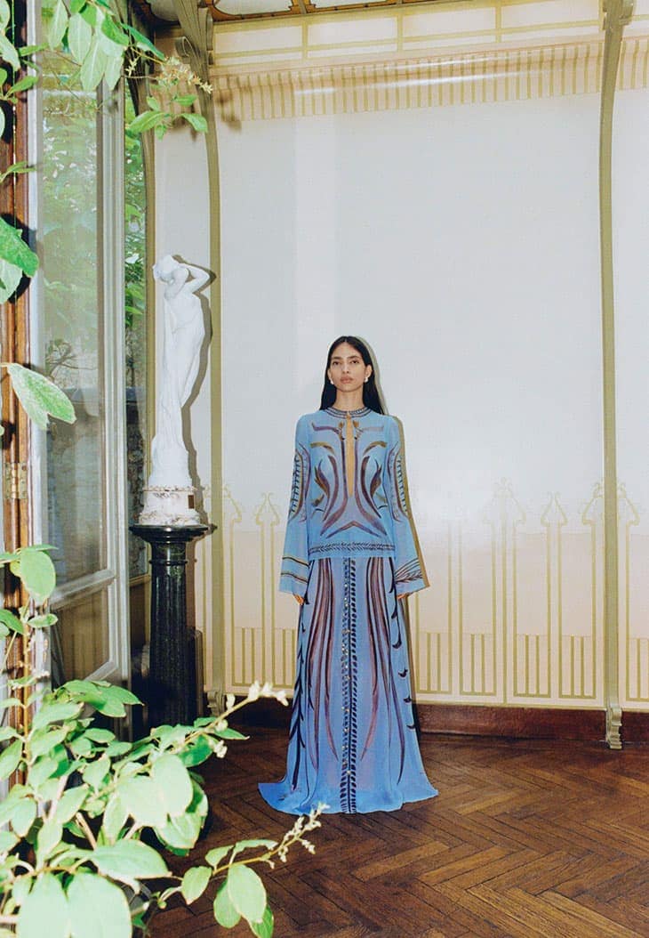 A fashionable adult female stands indoors wearing a long blue gown with brown patterning, complemented by a historic interior background.