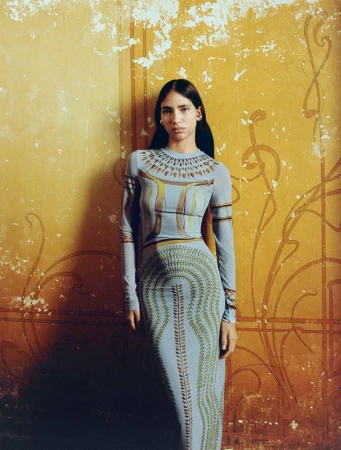 Adult woman poses in a patterned long-sleeved dress, standing indoors against a textured ochre wall with painted swirl details.