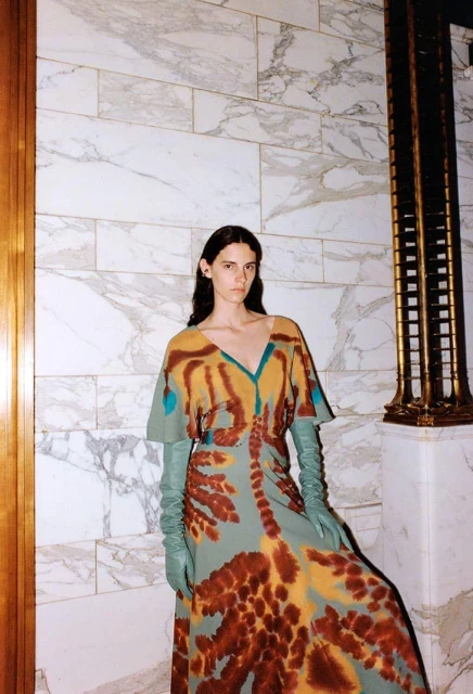 Elegant woman posing in a brown and turquoise patterned dress and long green leather gloves, standing against a marble wall.
