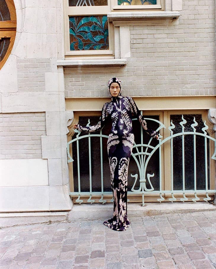 Full-length portrait of an adult woman in a patterned dress and head covering posing outdoors in front of a light-stone building.