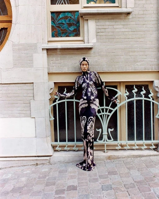 Full-length portrait of an adult woman in a patterned dress and head covering posing outdoors in front of a light-stone building.
