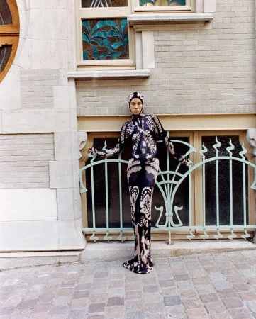 Full-length portrait of an adult woman in a patterned dress and head covering posing outdoors in front of a light-stone building.
