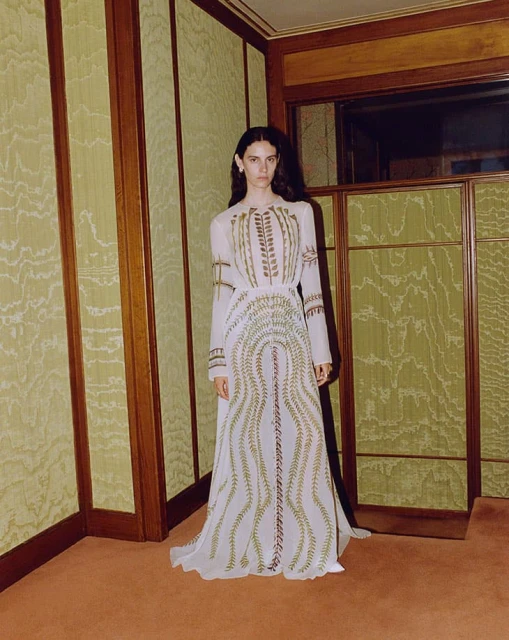 An adult female stands in an indoor setting with patterned wallpaper wearing a long white gown with decorative gold accents.