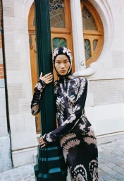 Model in printed long-sleeve dress and matching hood leaning against a dark green pole outdoors in Brussels