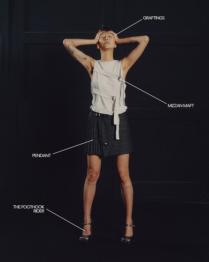 A slender, fair-skinned adult woman poses in a sleeveless white top, gray pleated skirt, and high heels against a dark background.