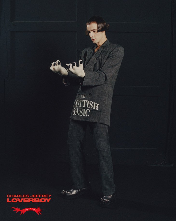 Man in grey suit with graphic, silver shoes poses in dark studio showcasing fashion and style, emphasizing outfit and modern design.
