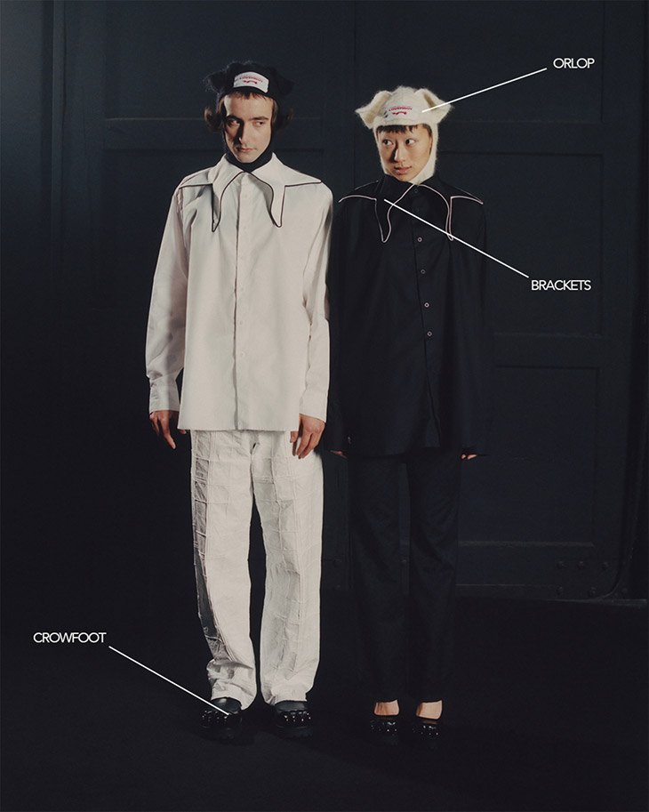 Two people stand in a photo studio wearing matching black and white outfits with unusual collars and hats, creating a symmetrical look.