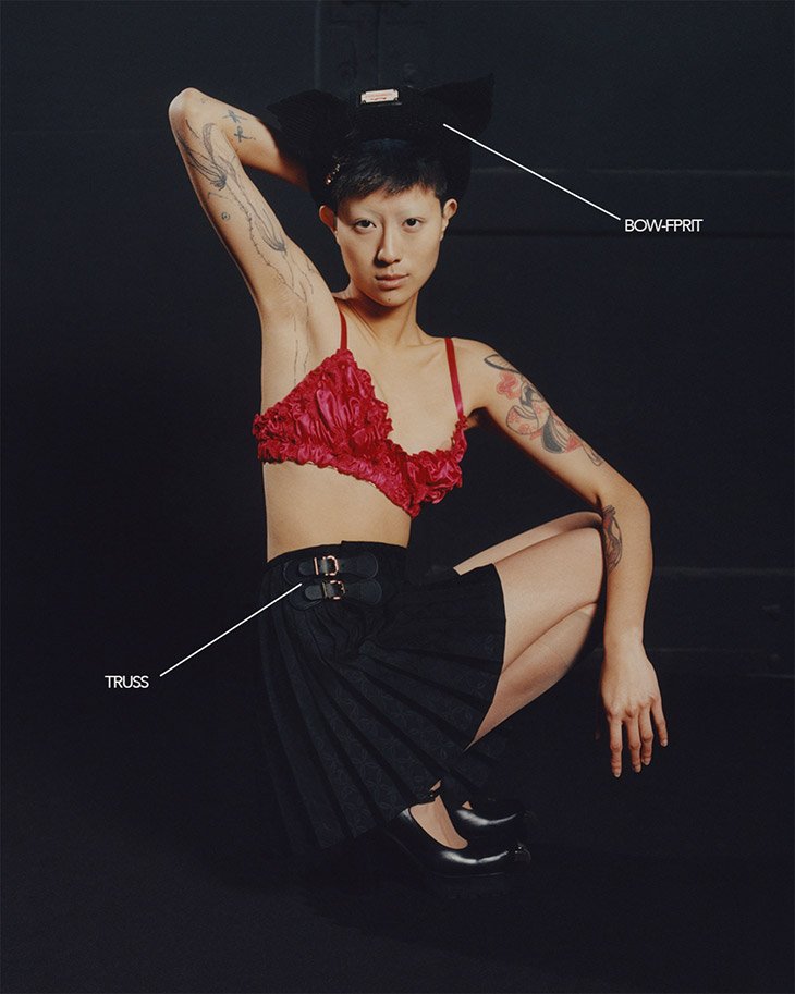 Asian female model squats in a photo studio wearing a red bra top, pleated mini-skirt, black flats and a sculptural black headpiece.