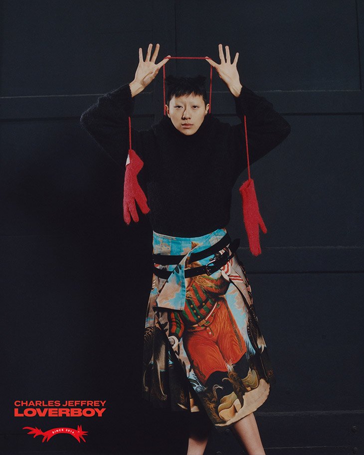 Fashion shot of person wearing a black turtleneck and a printed skirt, with red gloves hanging from a red string in a studio setting.