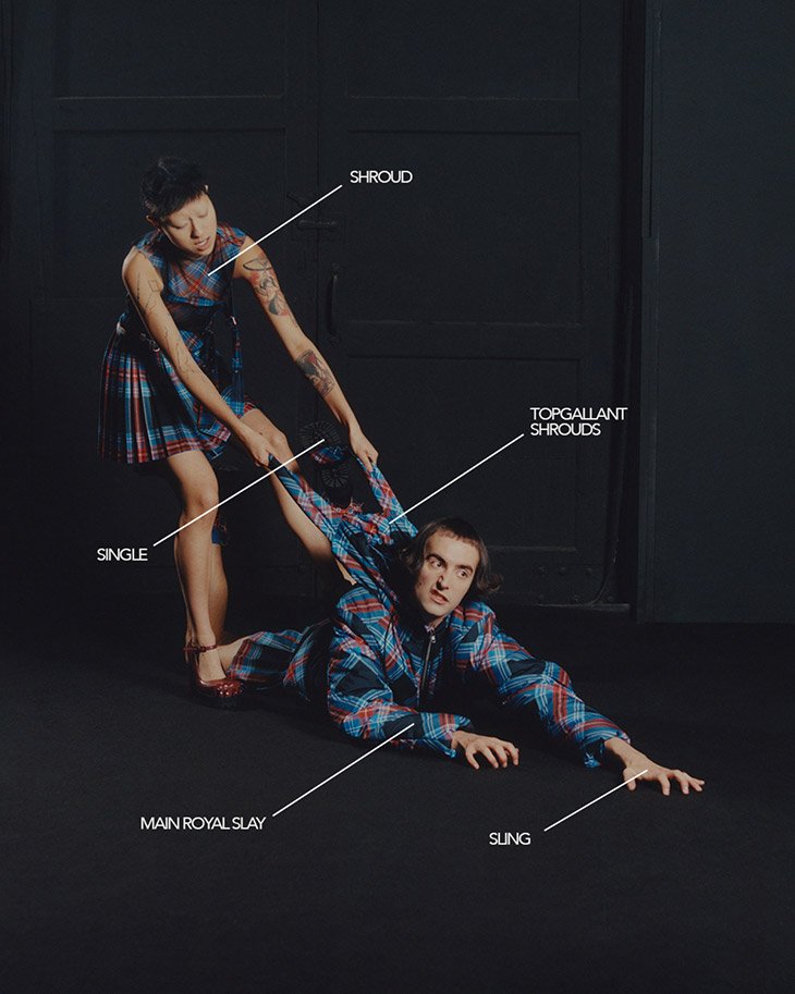 Two fashion models in a photo studio. An adult woman pulls on the tartan shirt of an adult man lying on the black studio floor.