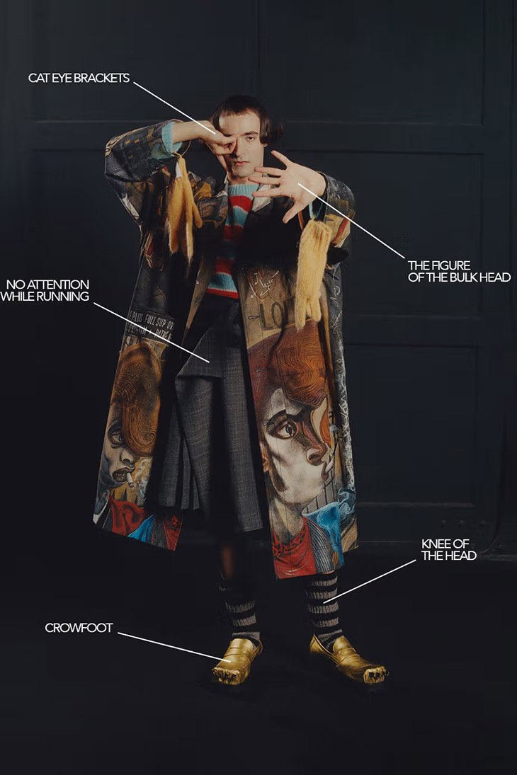 An adult male is standing in a studio wearing a graphic print coat over a grey pleated skirt with socks and gold shoes, posing expressively.