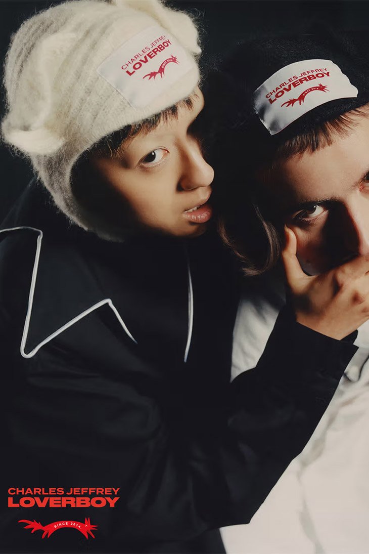 Two people pose in a dark studio wearing Charles Jeffrey Loverboy branded headwear and clothing with white detailing; studio lit fashion shot.
