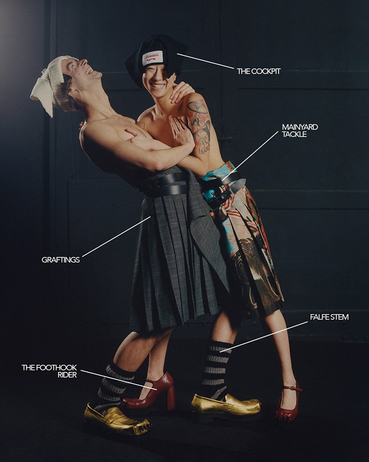 Two partially nude adults pose indoors, wearing skirts and socks, and striking a playful pose. One person has a white cloth on their head.