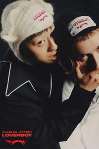 Two models wearing Charles Jeffrey Loverboy beanies and dark jackets posing closely against a dark studio background.