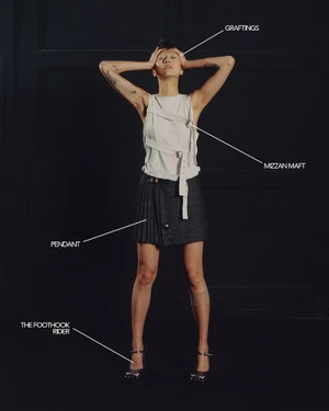 Model standing against dark background wearing white top and pleated skirt with diagrammatic callout text labels pointing to outfit elements.