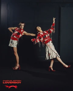 Two models in red patterned knitwear and printed skirts posing against a dark industrial background for Charles Jeffrey Loverboy